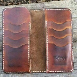 May include: Open brown leather wallet with multiple card slots and white stitching. The wallet has a worn, rustic appearance, with a natural leather texture and a stamped logo on the lower right side. The wallet is open, revealing the interior.