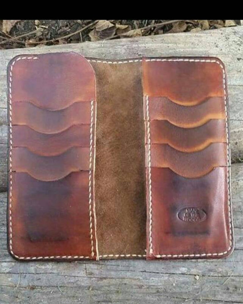 May include: Open brown leather wallet with multiple card slots and white stitching. The wallet has a worn, rustic appearance, with a natural leather texture and a stamped logo on the lower right side. The wallet is open, revealing the interior.