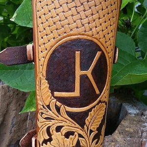 May include: A handcrafted leather arm guard in shades of brown, featuring a woven pattern at the top and floral tooling. A dark brown circle with the letter "K" is centered on the guard. The guard has a buckle closure.