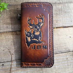May include: A brown leather wallet with a carved design of two deer and the initials "JDM". The wallet has a stitched border and leaf corner accents. The deer are depicted in black against the brown leather.