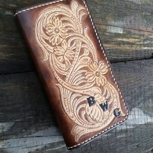 May include: A brown leather wallet with a floral design and the initials "BWG". The wallet features a detailed embossed pattern with a light tan color, and is stitched with white thread. The wallet is rectangular and appears to be closed.