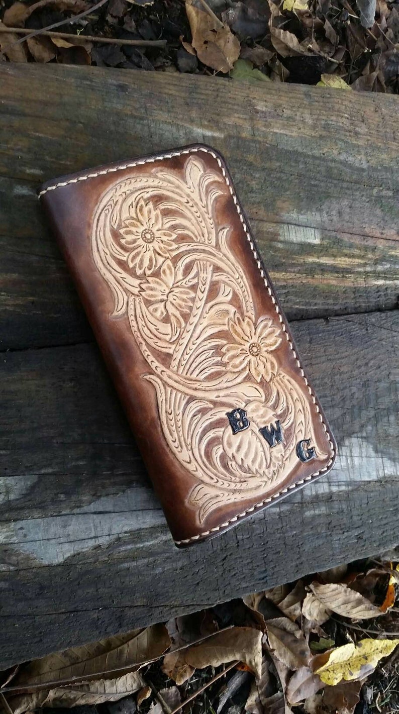 May include: A brown leather wallet with a floral design and the initials "BWG". The wallet features a detailed embossed pattern with a light tan color, and is stitched with white thread. The wallet is rectangular and appears to be closed.