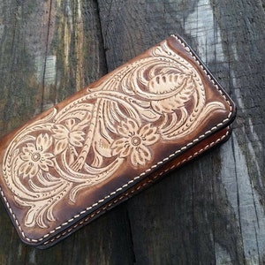 May include: A brown leather wallet with a floral design. The wallet is rectangular and features a detailed embossed pattern of flowers and leaves. The edges are stitched with white thread, and the leather has a gradient of light and dark brown tones.