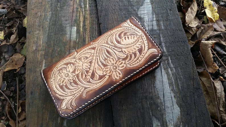 May include: A brown leather wallet with a floral design. The wallet is rectangular and features a detailed embossed pattern of flowers and leaves. The edges are stitched with white thread, and the leather has a gradient of light and dark brown tones.