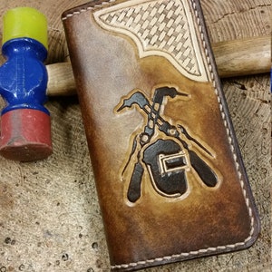 May include: A rectangular brown leather wallet featuring a hand-tooled design of crossed welding torches and a welding helmet. The top corner has a basketweave pattern, and the edges are finished with thick, light-colored stitching.