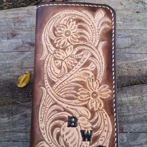 May include: A brown leather wallet with a floral design and the initials "BWG" embossed on the front. The wallet features a detailed carved pattern and is stitched with a lighter thread. The background is a wooden surface.