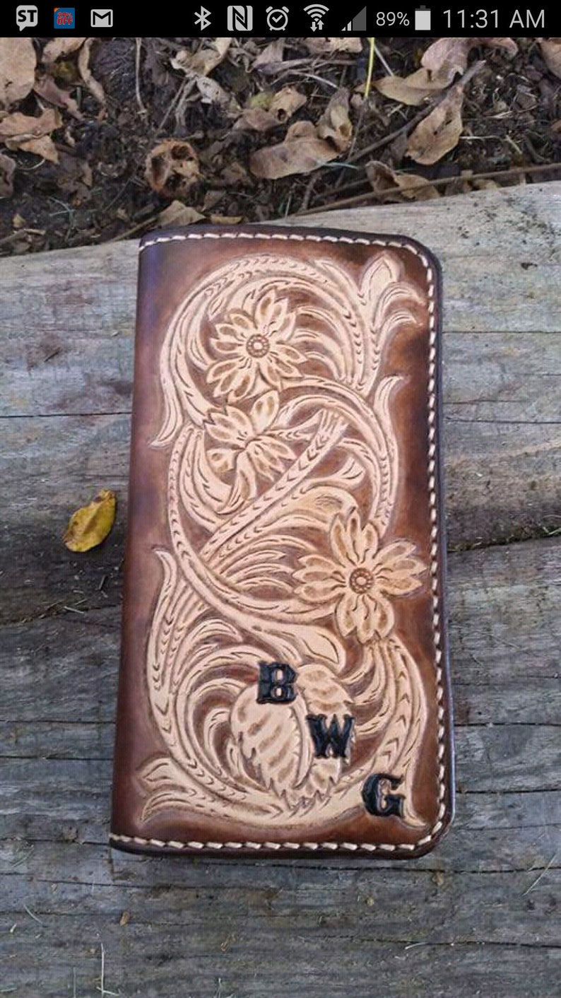 May include: A brown leather wallet with a floral design and the initials "BWG" embossed on the front. The wallet features a detailed carved pattern and is stitched with a lighter thread. The background is a wooden surface.