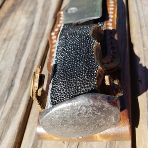 May include: A black and silver knife with a leather sheath. The handle is covered in a black material that resembles fish scales.