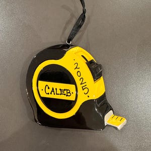 May include: A black and yellow tape measure ornament with the name "CALEB" and the year "2025" written on it. The ornament has a black ribbon for hanging.