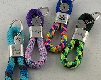 May include: Four braided rope keychains with silver metal clasps and circular charms. The keychains feature blue, purple, rainbow, and green patterns. Each metal clasp is engraved with the text Schulkind and a small icon, while the circular charms are engraved with the letters M, S, F, and A.