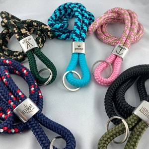 Könnte beinhalten: Sechs bunte Schlüsselbänder aus Seil mit Metallringen und gravierten Metalletiketten. Die Etiketten haben verschiedene Sprüche, darunter "Glück Auf", "Prinz", "Moin", "Heimat Hafen", "Lieblings Kollegin".