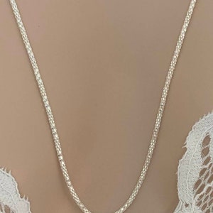 May include: A delicate silver necklace with a textured, beaded chain. The necklace is displayed against a neutral background, highlighting its intricate design. The chain's texture catches the light, creating a subtle sparkle. A simple and elegant piece of jewelry.