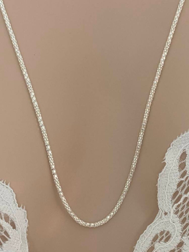 May include: A delicate silver necklace with a textured, beaded chain. The necklace is displayed against a neutral background, highlighting its intricate design. The chain's texture catches the light, creating a subtle sparkle. A simple and elegant piece of jewelry.
