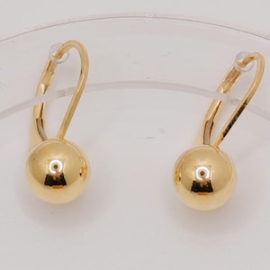 Peut inclure: Une paire de boucles d'oreilles en or présentant un design en forme de boule sphérique polie suspendue à des crochets incurvés. La surface lisse et réfléchissante des sphères dorées capte la lumière, offrant une esthétique simple et épurée.