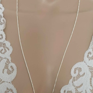 Peut inclure: Un délicat collier en chaîne argentée. La chaîne présente un motif tissé texturé et pend sur un fond neutre. Le collier est présenté sur un vêtement en dentelle blanche.