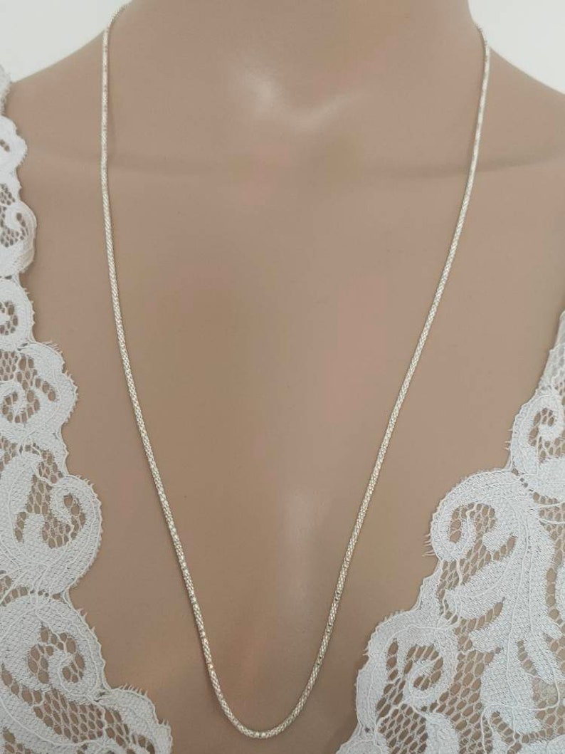 May include: A delicate silver chain necklace. The chain has a textured, woven design and hangs down against a neutral background. The necklace is displayed against a white lace garment.