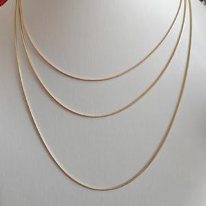 Op de afbeelding: Een set van drie goudkleurige slangenkettingen van verschillende lengtes, gelaagd om een waterval-effect te creëren. Elke dunne, flexibele ketting heeft een glad, gepolijst oppervlak dat licht reflecteert voor een uniforme, metalen uitstraling.