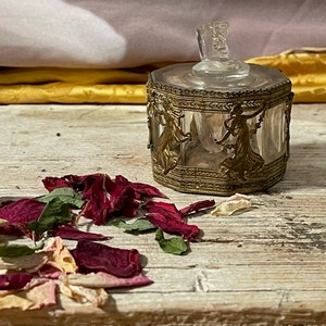 May include: A small, clear glass jar with a gold metal lid and decorative trim. The jar is filled with dried rose petals and is sitting on a wooden surface.