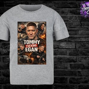May include: A light gray t-shirt featuring a graphic of a man with the text "TOMMY FUCKING EGAN". The graphic includes images of the man in various poses. The shirt has a crew neck and short sleeves.