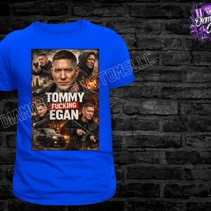 May include: A royal blue t-shirt featuring a graphic of a man with the text "TOMMY FUCKING EGAN". The graphic includes images of other men and a car. The shirt has a crew neck and short sleeves.