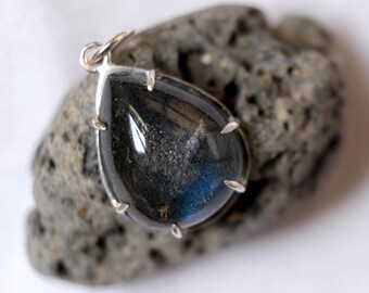 May include: A teardrop-shaped pendant with a silver setting, showcasing a dark, iridescent gemstone with blue and gray hues. The pendant is resting on a rough, gray stone, creating a contrast in textures. A jewelry item.