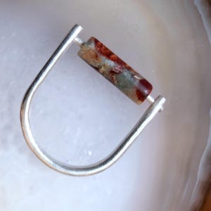 May include: A silver ring with a rectangular, multi-coloured stone. The stone shows shades of red, brown, and green. The ring has a simple, modern design, with a curved band.