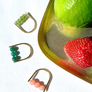 Op de afbeelding: Drie U-vormige ringen van goudkleurig metaal, elk voorzien van een rij kleine kralen. De bovenste ring heeft vier lichtgroene gefacetteerde kralen, de middelste ring heeft drie gladde donkergroene kralen en de onderste ring heeft drie lichtroze kralen.