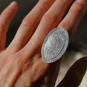 May include: A silver ring with an oval, textured face. The ring is worn on a finger, showcasing its detailed design. The ring's surface has a brushed metal appearance, reflecting light. The ring is a piece of jewelry.