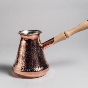 Puede incluir: Una cafetera de cobre con acabado martillado y mango de madera. La cafetera está diseñada para preparar café turco.