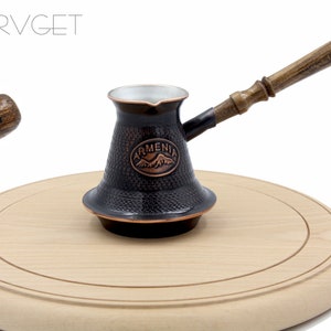 May include: A black and copper Turkish coffee pot with a wooden handle. The pot has a textured surface and the words "Armenia" and a mountain range are embossed on the side. The pot is sitting on a wooden cutting board.