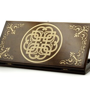May include: A wooden game box with a brown finish and intricate gold-colored designs. The box is closed and features a circular design with a knot pattern in the center.