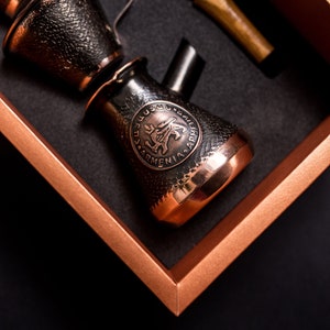 Puede incluir: Una cafetera de cobre con un acabado texturizado negro y un diseño circular con un pájaro estilizado y el texto "ARMENIA". La cafetera está en una caja marrón con un interior negro.