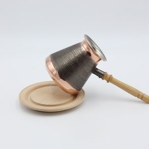 May include: A copper and stainless steel coffee pot with a hammered finish and a wooden handle. The pot is sitting on a wooden coaster.