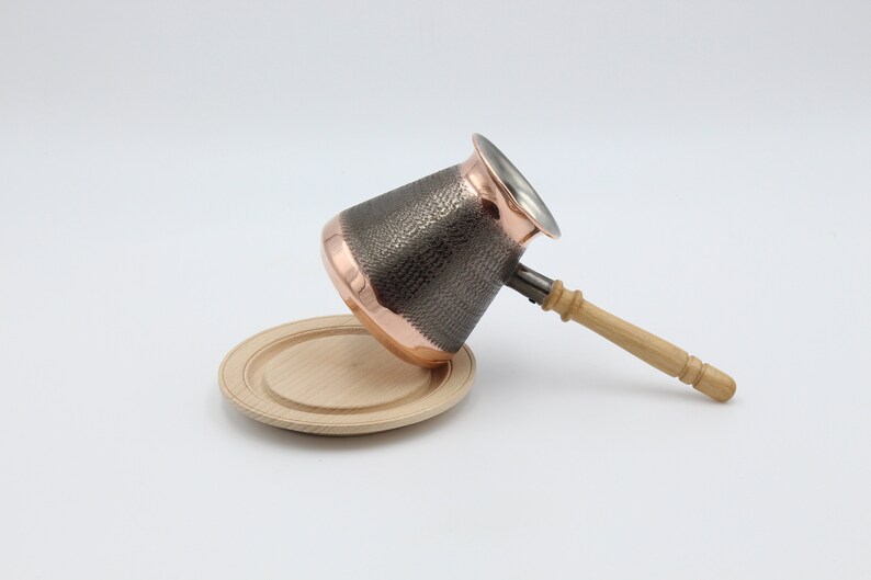 May include: A copper and stainless steel coffee pot with a hammered finish and a wooden handle. The pot is sitting on a wooden coaster.