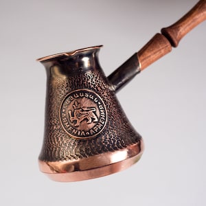 Op de afbeelding: Een koperen koffiepot met een houten handvat. De pot heeft een gehamerde textuur en heeft een rond embleem met het Armeense wapen en de woorden "ՀԱՅԱՍՏԱՆ" en "ARMENIA" rond de rand.