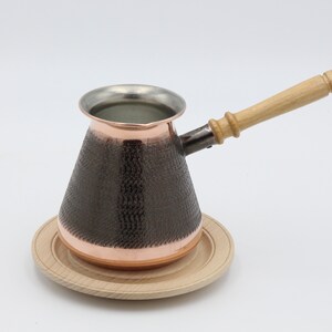 May include: A copper coffee pot with a hammered finish and a wooden handle. The pot is sitting on a wooden coaster.
