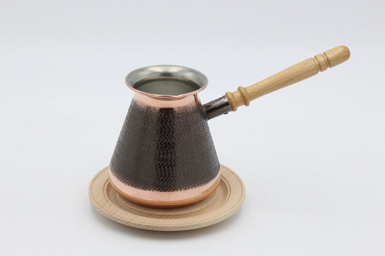 May include: A copper coffee pot with a hammered finish and a wooden handle. The pot is sitting on a wooden coaster.