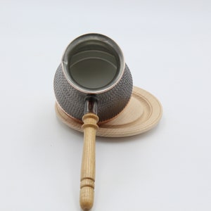 May include: A copper and black coffee pot with a wooden handle, sitting on a wooden coaster.