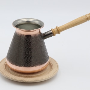 May include: A copper coffee pot with a wooden handle and a textured surface. The pot is sitting on a wooden coaster.