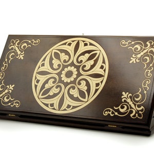 May include: A dark brown wooden backgammon board featuring a central, light-coloured floral design. The board has decorative corner detailing and is partially open, revealing the game's interior. The board's dimensions are in centimetres.