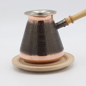 May include: A copper Turkish coffee pot with a wooden handle and a hammered finish. The pot is sitting on a wooden coaster.