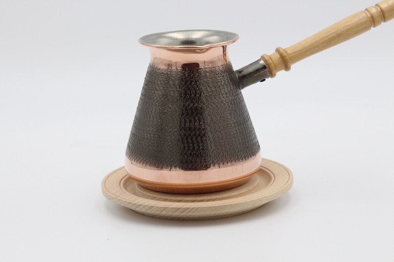 May include: A copper Turkish coffee pot with a wooden handle and a hammered finish. The pot is sitting on a wooden coaster.