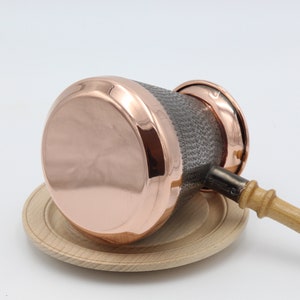 May include: A copper coffee pot with a wooden handle and a hammered texture. The pot is sitting on a wooden base.