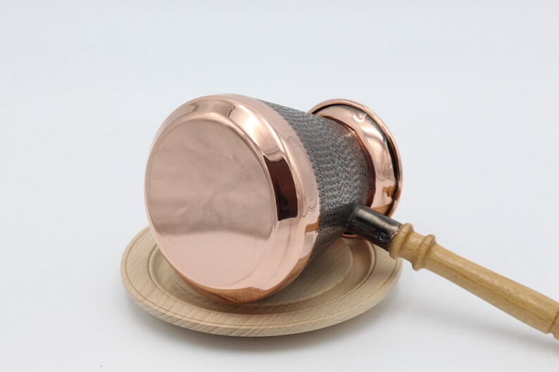 May include: A copper coffee pot with a wooden handle and a hammered texture. The pot is sitting on a wooden base.