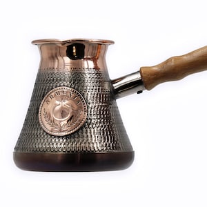 Può includere: Una caffettiera in rame e argento con un manico in legno. La caffettiera ha una finitura martellata e un emblema rotondo con la parola "Armenia" e un motivo di melograno.
