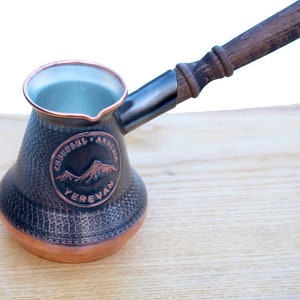 Puede incluir: Una cafetera de cobre y negro con un mango de madera. La cafetera tiene una superficie texturizada y un logotipo circular que dice "Cakusuli Armenia Yerevan".
