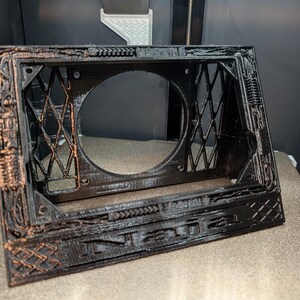 May include: A black 3D printed frame with copper-colored highlights featuring a central circular opening, a diamond-patterned lattice on the sides, and the word "Naja" embossed in capital letters along the bottom front edge of the structure.