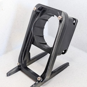 Puede incluir: Un soporte de ventilador negro impreso en 3D con una abertura circular y patas de soporte anguladas. El soporte se asegura con pequeños tornillos plateados. El diseño parece ser para un pequeño dispositivo o componente electrónico.