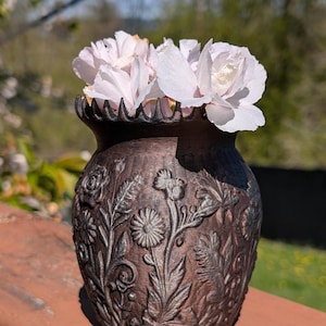 May include: A bronze-colored vase with a detailed floral relief design, filled with delicate, light pink flowers. The vase has a textured surface and a scalloped rim. The flowers are in full bloom, adding a soft contrast to the vase's darker tones.