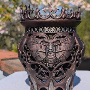 May include: A bronze-colored decorative vase with a crown-like top and intricate cut-out designs. The vase features a moth with a skull on its body. The vase is approximately 6 inches tall.